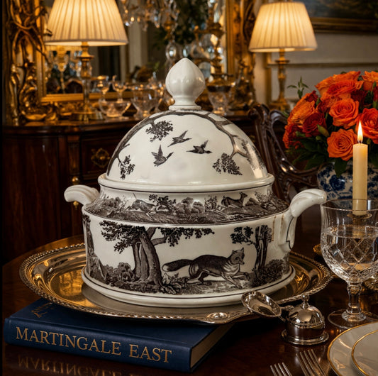 Decorative white tureen with black animal illustrations on a table with books and glasses.