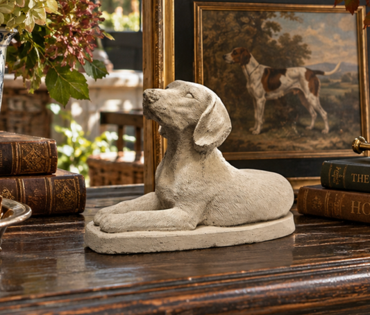 A light beige stone statue of a lying dog, displayed on a dark wooden table in a styled room setting with antique books and a framed painting of a hunting dog.
