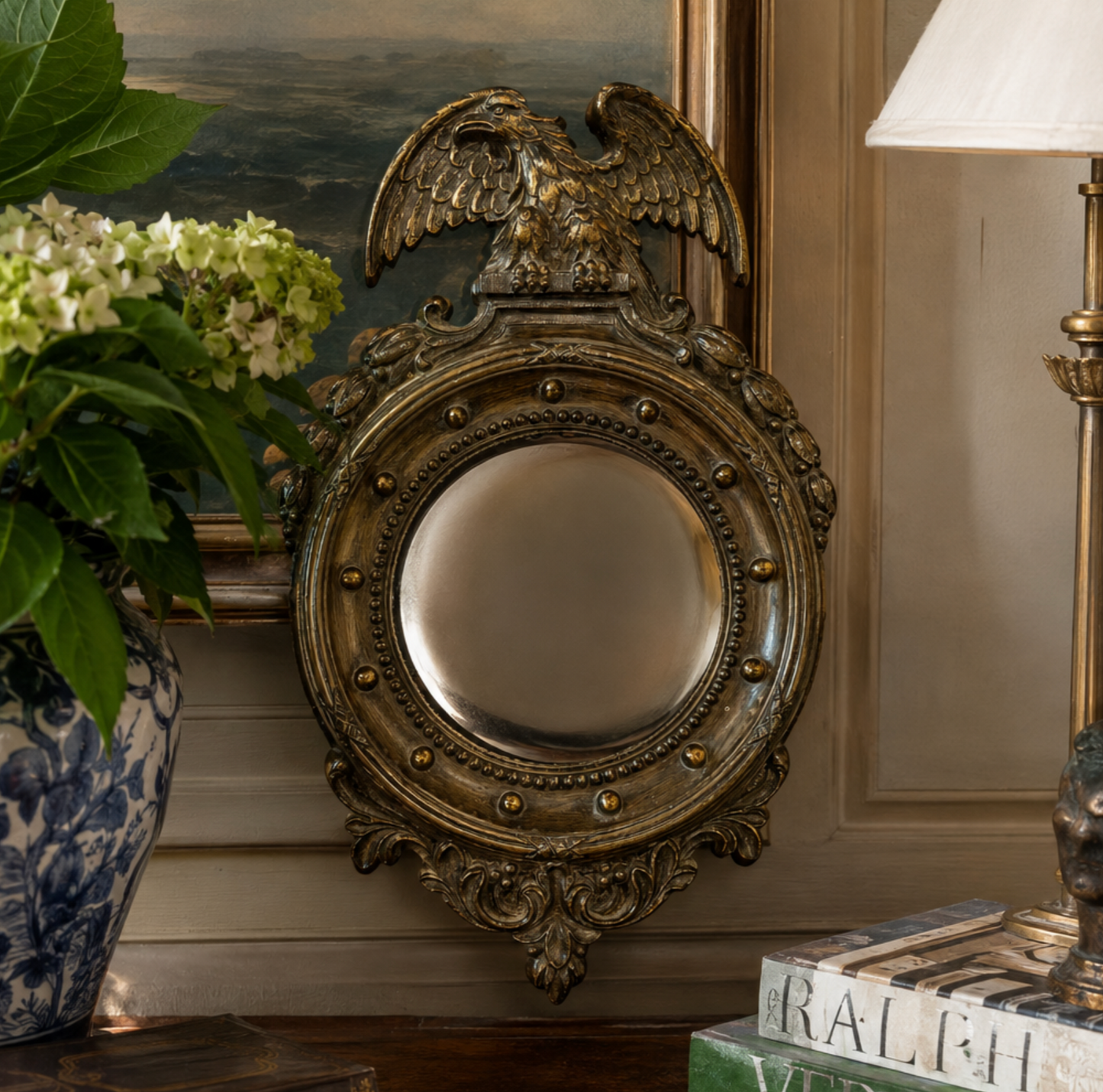 An ornate antique-style round wall mirror with a bronze eagle crest and decorative scrollwork frame, displayed in a styled interior setting next to a blue-and-white vase and a table lamp.