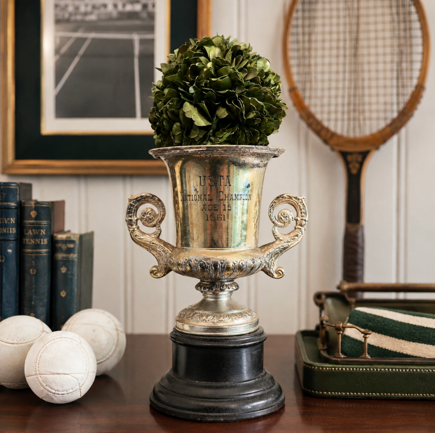 A decorative silver trophy cup with a black pedestal base holding a green leafy plant, styled on a wooden table with tennis-themed props including a racket, books, balls, and a case in an indoor room setting.