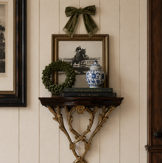 A decorative wall-mounted console shelf in a warm wood finish with ornate gold-toned bracket legs, styled with a black-and-white equestrian photo print in a gold frame, a green wreath, a blue-and-white porcelain jar, and books against a cream paneled wall.