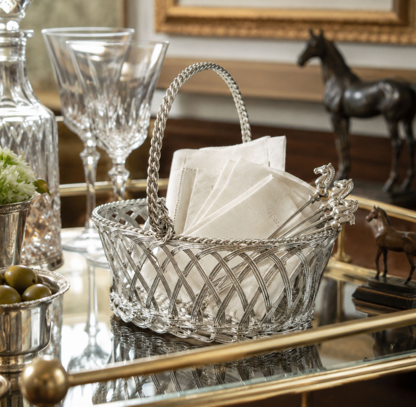 A silver wire basket with a decorative handle and silver serving utensils, filled with white napkins, displayed on a glass tray in an elegant interior setting.